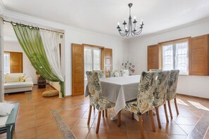Un comedor con una mesa dispuesta para cuatro, sillas con estampado floral, una araña y vistas a través de una ventana con contraventanas de madera.