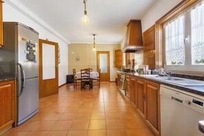 Een keuken met een koelkast, vaatwasser, houten keukenkastjes, een eettafel met stoelen en een raam met gordijnen.