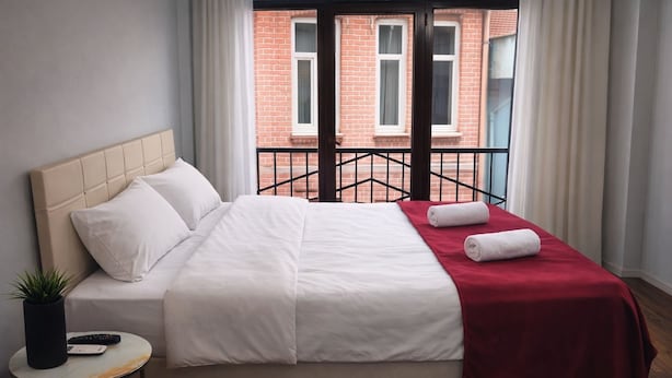 A neatly made bed with white and red bedding, a small round table with a plant and a remote control, and a view of a red brick building through a window with a black metal railing. - Balatlifesuites (İstanbul)