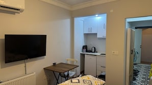 A compact room with a flat-screen TV, a small table, a chair, a kitchenette with cabinets, a coffee maker, and a patterned rug. - Balatlifesuites (İstanbul)