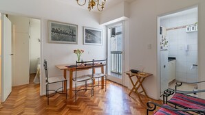 A room with a wooden herringbone floor, a dining table with chairs, a small side table, a bed with a red and blue striped bedspread, a chandelier, and a kitchen area with a refrigerator and cabinets. - Warm and cozy apartment for two in Palermo (CABA)