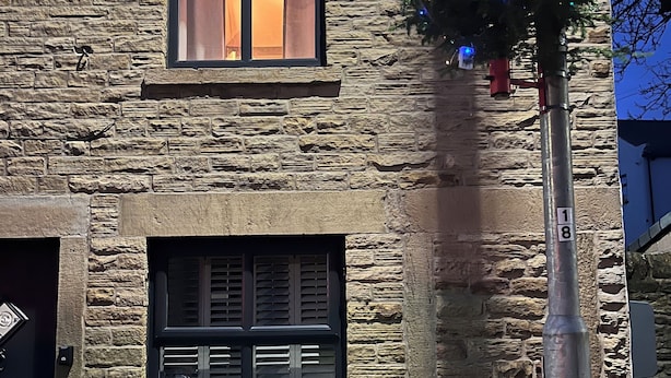 Un bâtiment en pierre doté de deux fenêtres, dont l’une est pourvue de volets et l’autre d’un rideau.
