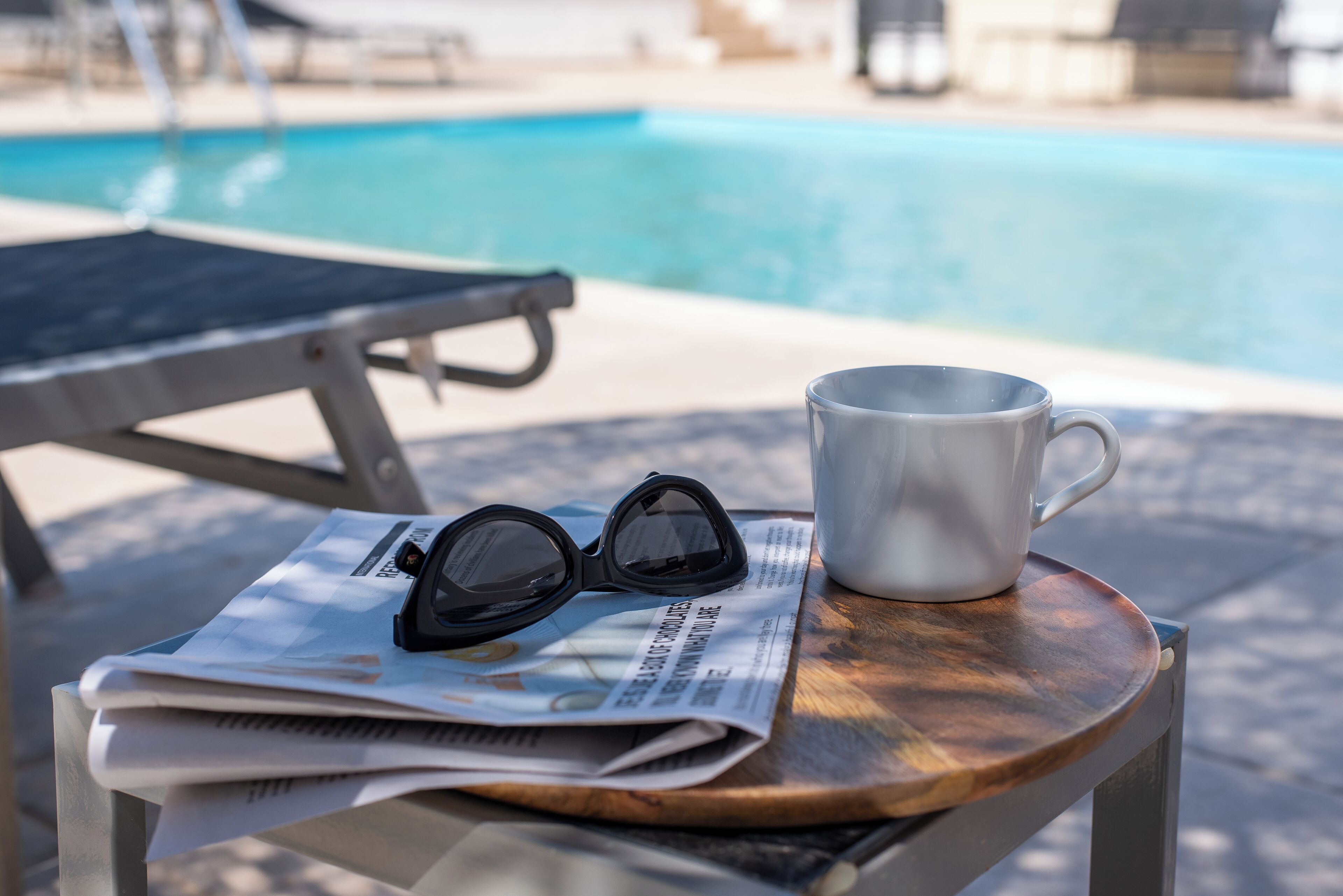 A newspaper, sunglasses, and a mug on a wooden table by a poolside.