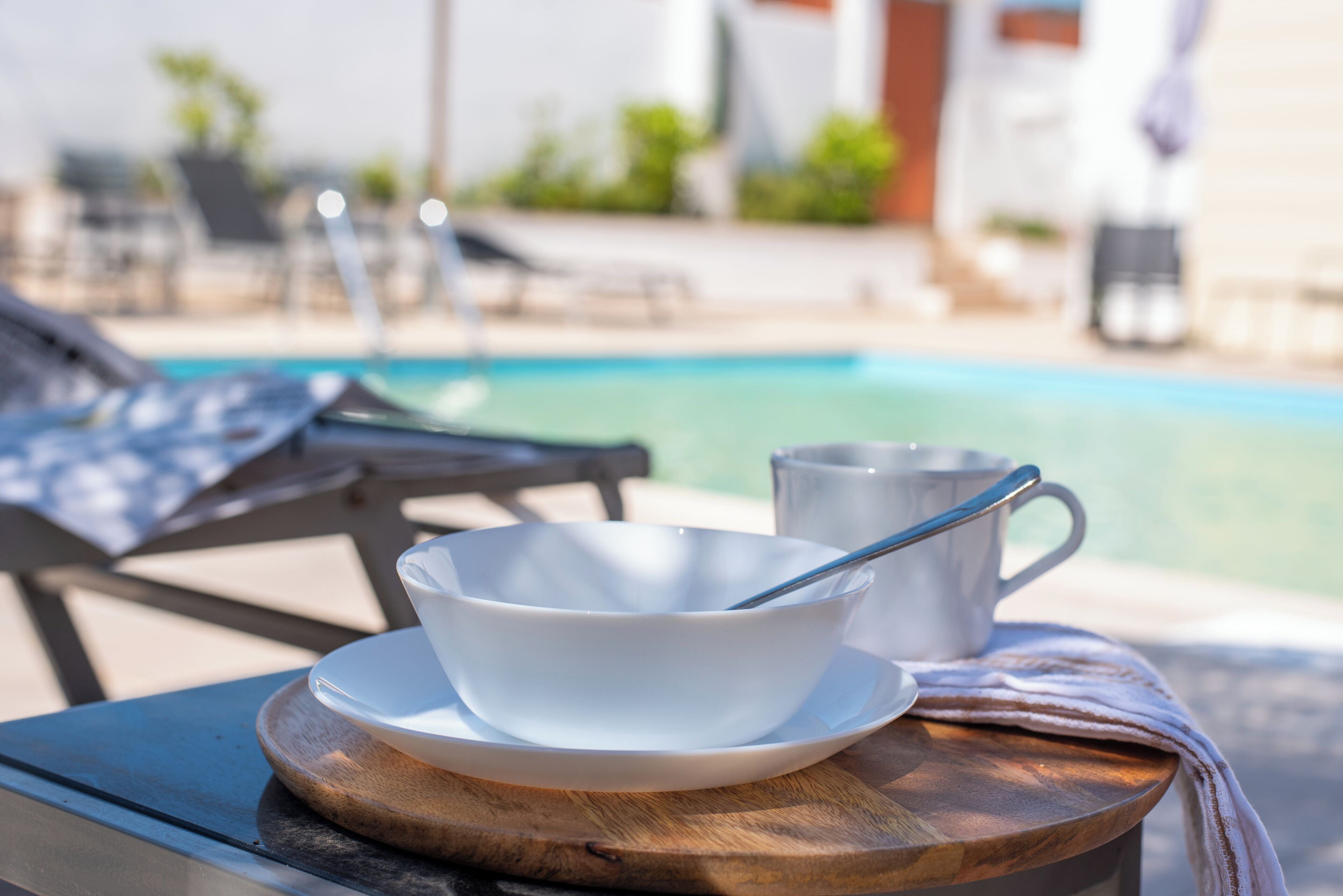 A white bowl and mug on a wooden table by a poolside.