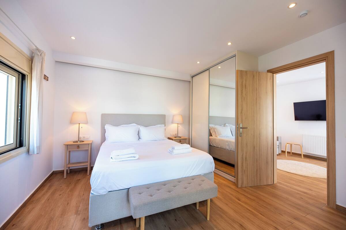 Ein modernes Schlafzimmer mit einem großen Bett, Nachttischlampen, einer Bank, einem Spiegel und einem Fernseher.