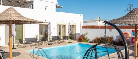 Une piscine entourée de chaises longues et de parasols, elle-même bordée d’un bâtiment blanc doté de grandes fenêtres et de grandes portes.