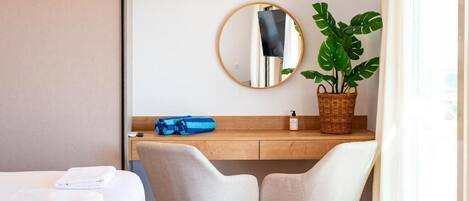 A hotel room with a bed, two white chairs, a wooden desk, a mirror, a potted plant, and a window with curtains.