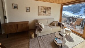 Een gezellige woonkamer met een houten plafond, een beige bank, een houten bijzettafeltje en een groot raam met uitzicht op een besneeerd landschap.