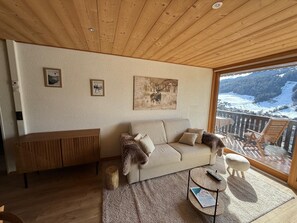 A cozy living room with a wooden ceiling, a beige sofa, a wooden side table, and a large window overlooking a snowy landscape. - Morz'Inn- Wake up to the mountains in Morzine (Morzine)