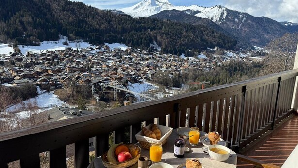 Mesa posta com itens de café da manhã, incluindo frutas, suco e doces, com vista para uma aldeia coberta de neve e montanhas.