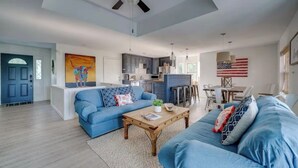 Ein geräumiges Wohnzimmer mit einem blauen Sofa, einem Holztisch und einem Deckenventilator. Im Hintergrund ist eine Küche mit modernen Geräten sowie ein Essbereich mit Tisch und Stühlen zu sehen.