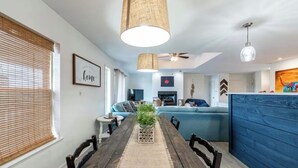 Ein Essbereich mit einem Holztisch, Stühlen und einer Zimmerpflanze. Im Hintergrund befindet sich eine blaue Couch und ein Deckenventilator.