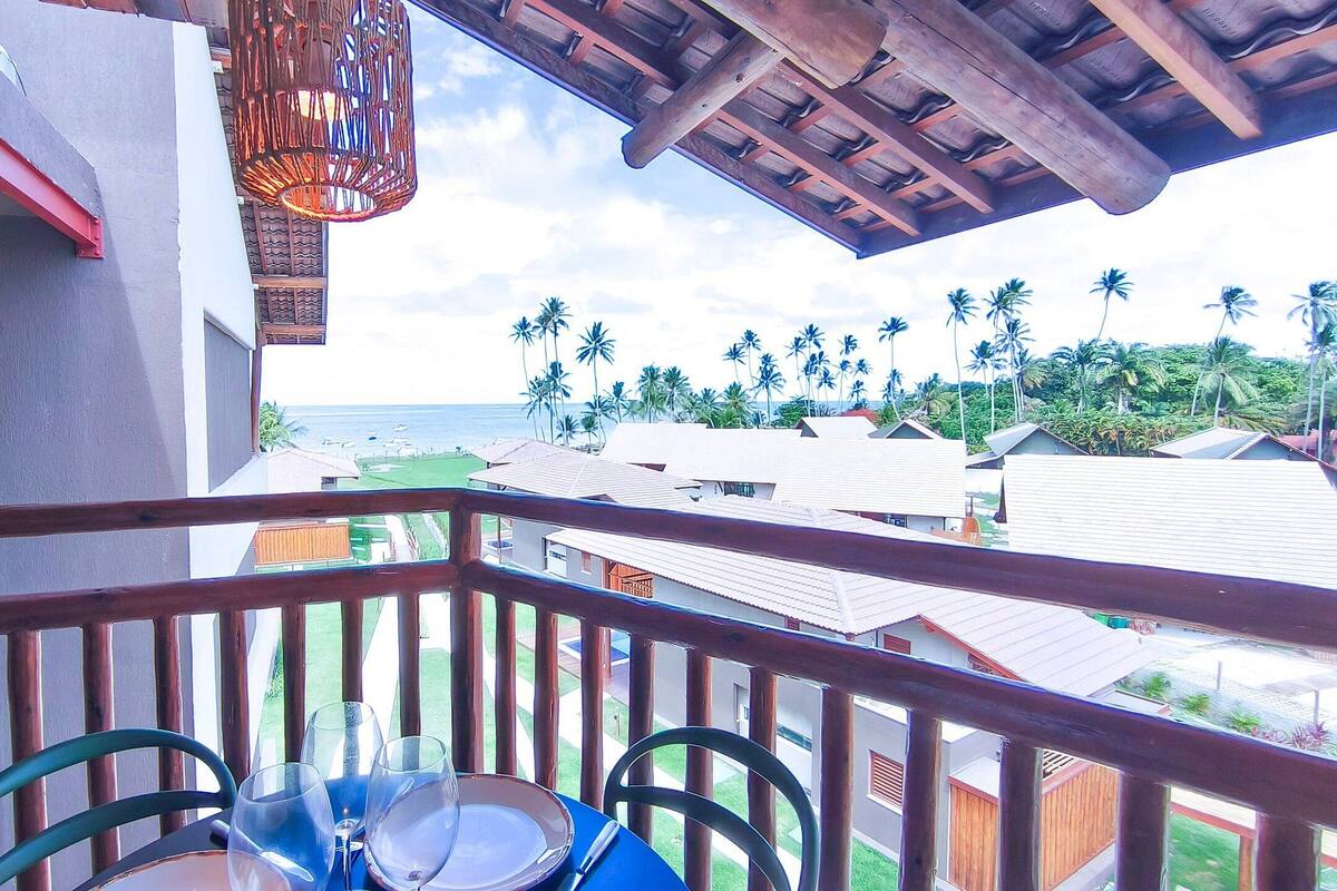 A balcony with a table set for dining, overlooking a residential area with houses and palm trees, and a view of the ocean.