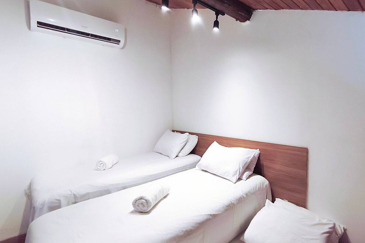Una camera d'albergo con due letti singoli, una testiera in legno, lenzuola bianche, un condizionatore d'aria e un lampadario.