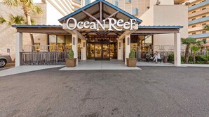 Der Eingang des Ocean Reef Hotels mit überdachtem Bereich, einer Palme und einer Person, die auf einer Bank sitzt.