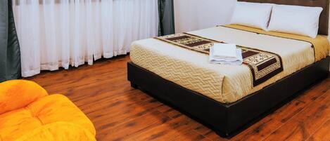 Habitación de hotel con cama, un ventanal con cortinas translúcidas, una lámpara de techo brillante y suelo de madera.