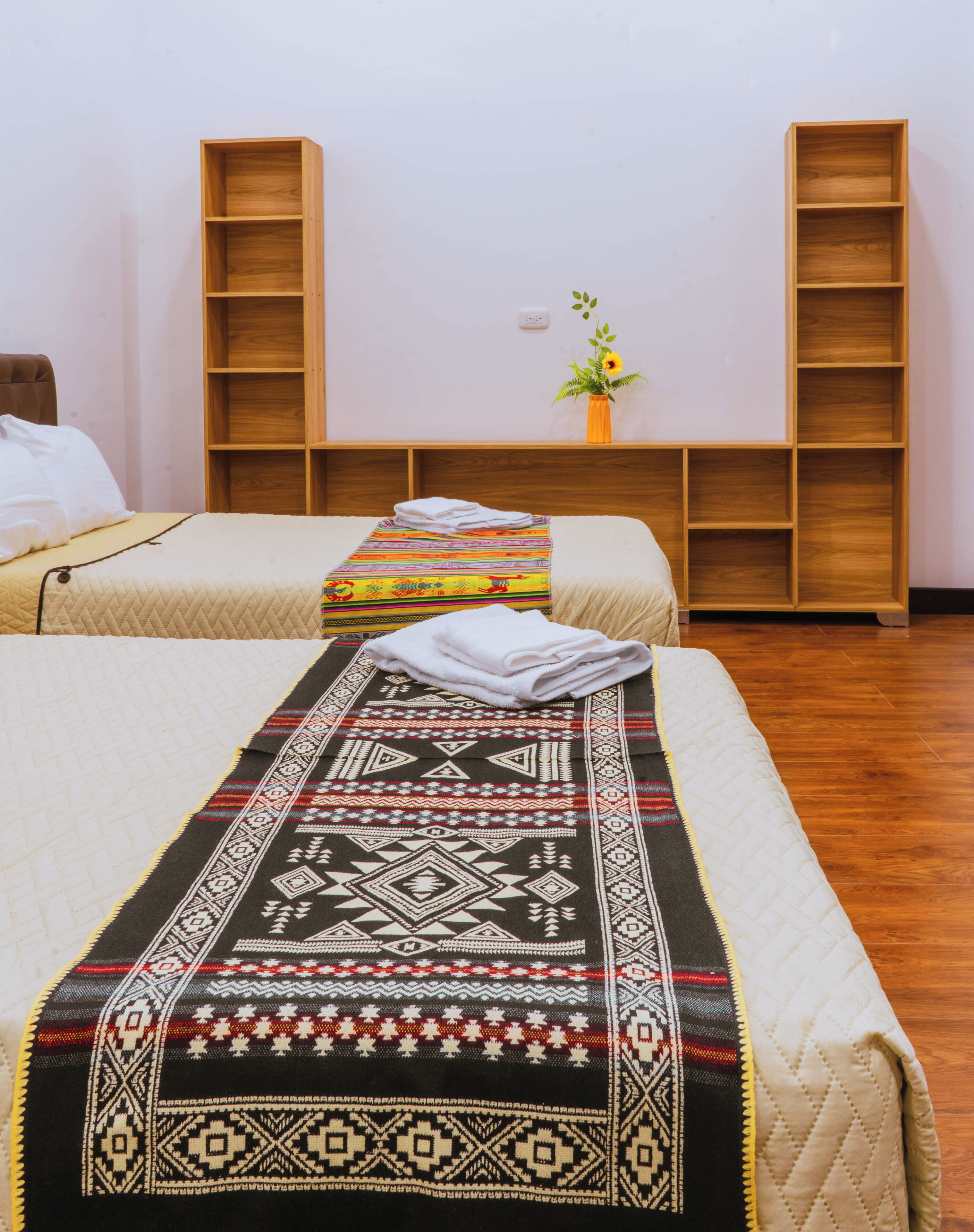 A hotel room with two beds, a wooden wardrobe, a small table with a vase, and a wall socket.