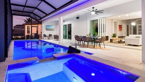 A modern indoor pool area with a glass ceiling, a seating area with chairs and a table, and a view of the outside. - Intervillas Florida - Villa Carrera (Cape Coral)