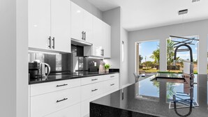 A modern kitchen with white cabinets, black countertops, and a large window offering a view of the outdoors. - Intervillas Florida - Villa Carrera (Cape Coral)