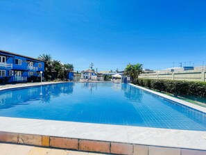 Uma piscina com superfície de água azul cristalina, rodeada por um muro baixo de tijolos e um edifício azul com grades brancas.