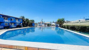 Una piscina azul cristalina rodeada de una terraza con baldosas, con un edificio azul y palmeras al fondo.