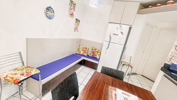 Una habitación pequeña y funcional con una cama individual, una mesa de madera, una silla, un refrigerador y un pequeño rincón de cocina.