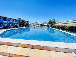 A clear blue swimming pool surrounded by a tiled deck, with a blue building and palm trees in the background. - Golden Dolphin (Porto Seguro)
