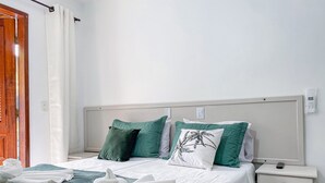 Een opgemaakt bed met een groene sprei en witte lakens, twee groene kussens, en een wit kussen met een palmboom dessin. Er liggen witte handdoeken op het bed en een witte nachtkastje met een afstandsbediening erop.