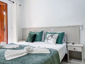 Un lit bien fait, recouvert d’une couvre-lit verte et de draps blancs, avec deux oreillers verts et un oreiller blanc orné d’un motif de palmier. Des serviettes blanches sont pliées sur le lit, et une table de chevet blanche, sur laquelle se trouve une télécommande, est placée à côté du lit.