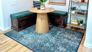 Ein gemütlicher Raum mit einem runden Holztisch, einer Bank mit einem grünen Kissen, einem blau gemusterten Teppich, Holzdielen, einem Fenster mit Jalousien, einem an der Wand befestigten Regal mit Pflanzen und Büchern sowie einer Deckenleuchte.