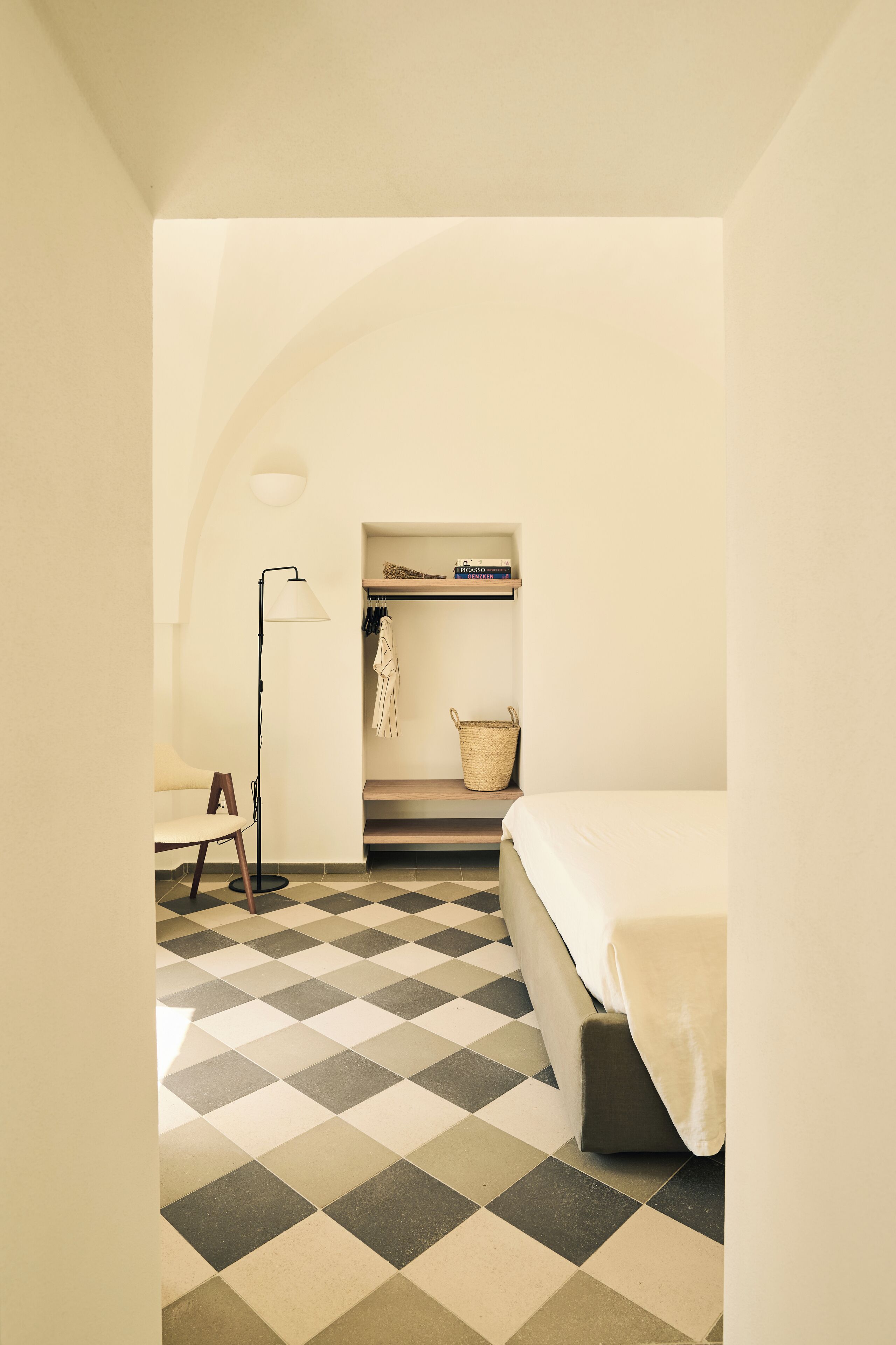 A compact room with a checkered floor, a bed, a desk with a chair, a floor lamp, a shelf with books, and a hanging towel.