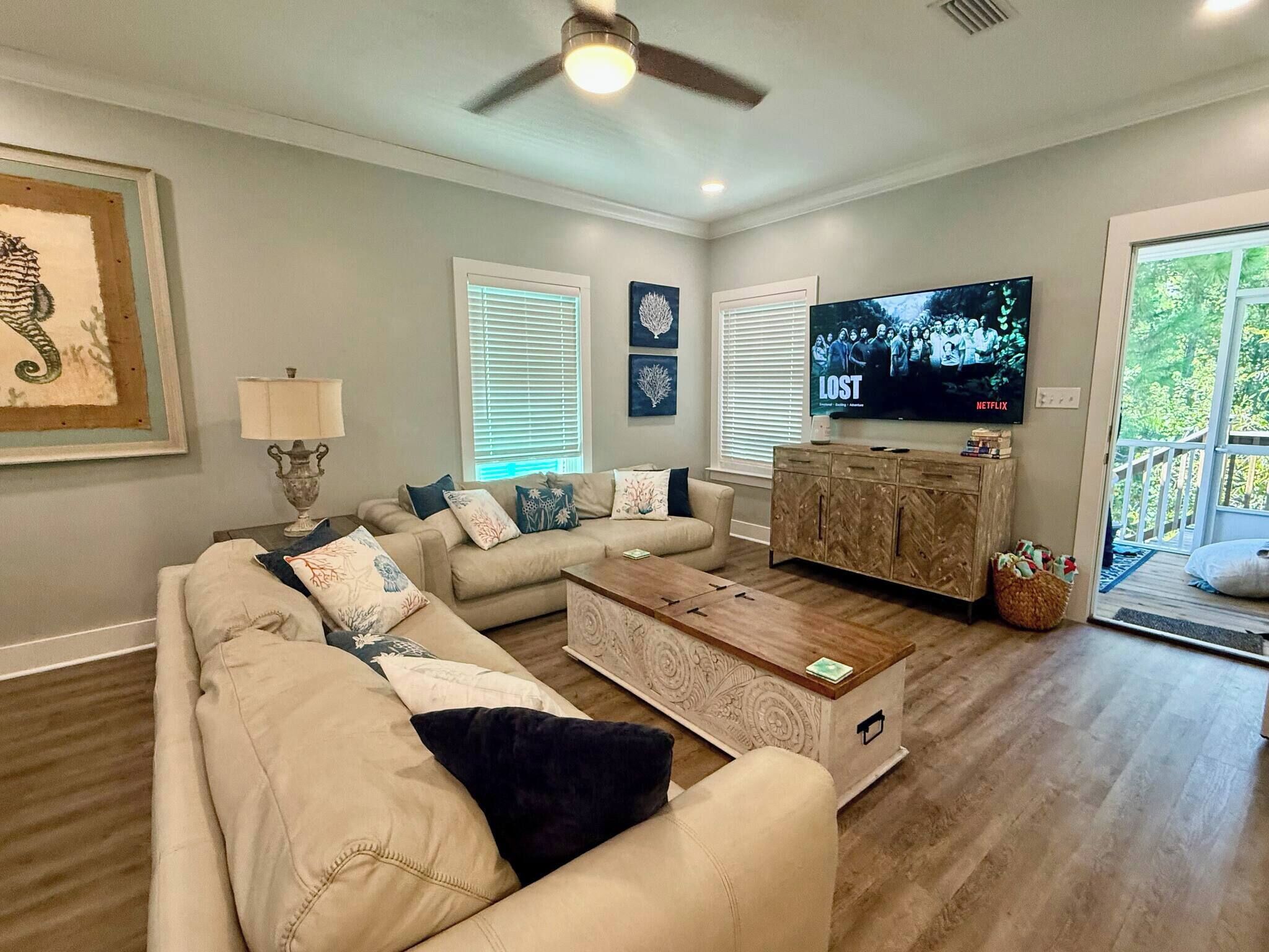 A living room with a beige sectional sofa, a wooden coffee table, and a TV on a wooden cabinet. There are two windows with blinds, a ceiling fan, and a framed picture on the wall.