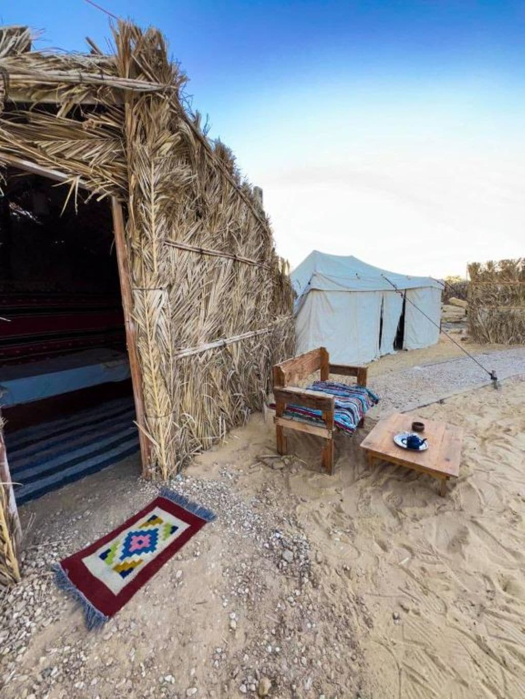 Un décor extérieur rustique avec une structure recouverte de chaume, une chaise en bois, une petite table avec une théière et une tente en arrière-plan.