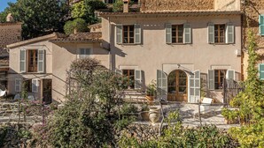 Un edificio de estilo mediterráneo tradicional con múltiples ventanas y contraventanas, rodeado de exuberante vegetación y muros de piedra.