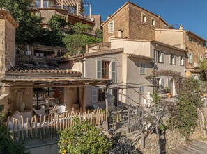 Ein traditionelles Steinhaus mit Balkon, Holzzaun und Gartenanlage mit Kübelpflanzen.