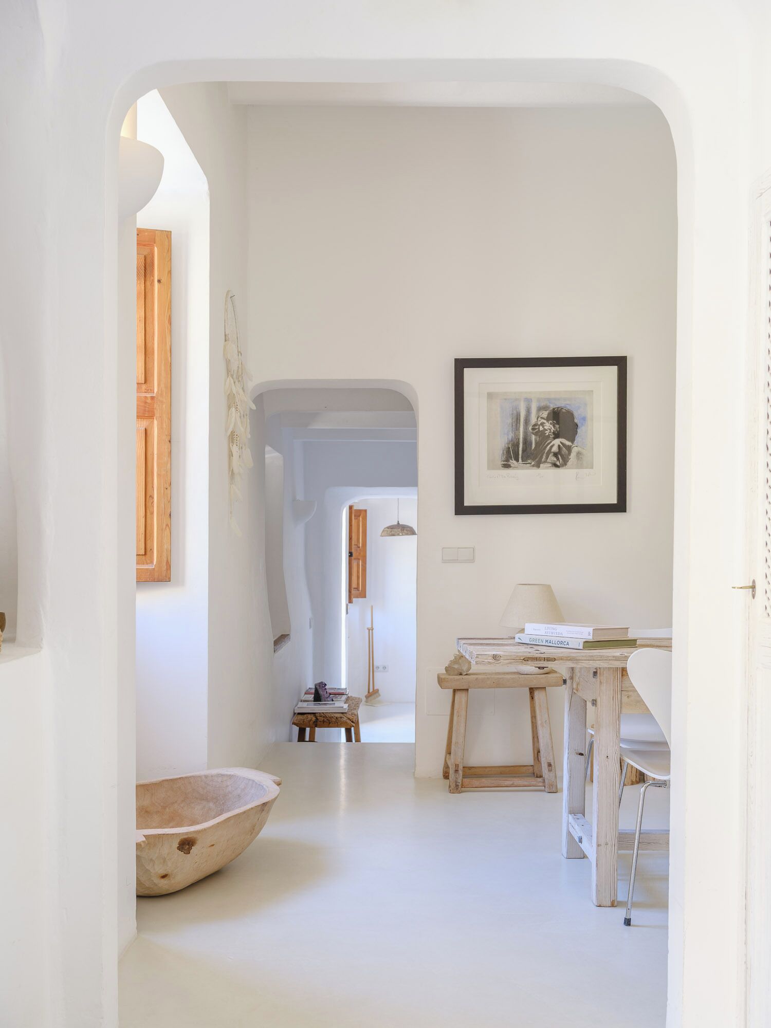 A minimalist interior with a white color scheme, a wooden table with books, a framed picture on the wall, and a large wooden bowl on the floor.