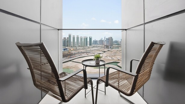 Un balcon avec deux fauteuils en osier, une petite table ornée d’une plante, et une vue sur un paysage urbain dominé par des immeubles de grande hauteur.