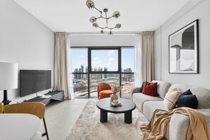 A modern living room with a large window, a flat-screen TV, a sofa, a coffee table, and a chandelier. - Silkhaus Radiant Bay (Abu Dhabi)
