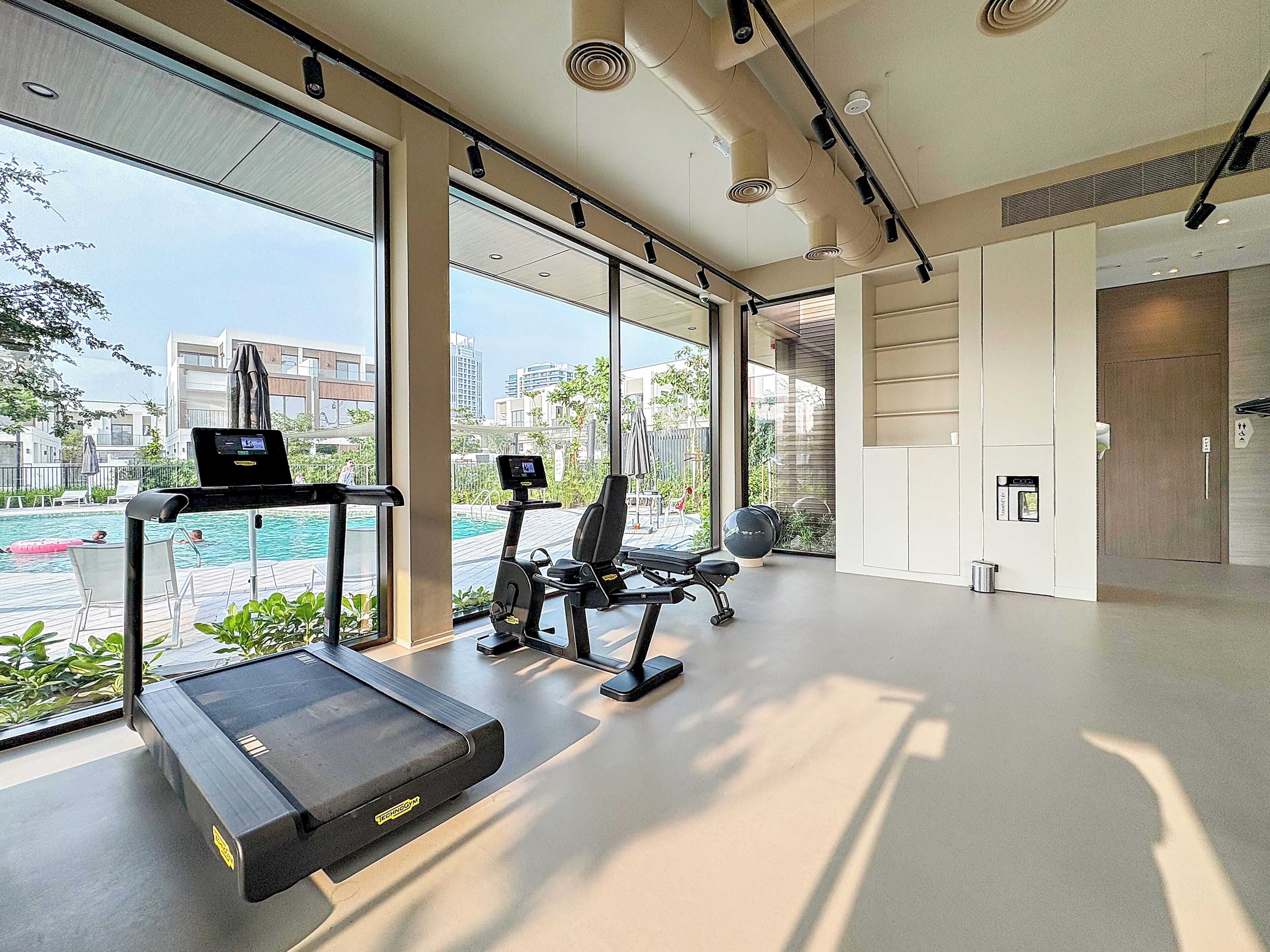 A modern gym with a treadmill, elliptical, and weight bench, large windows with a view of buildings and greenery.