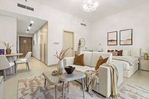Una sala moderna con un sofá, una mesa de comedor y una mesa de centro. Hay una cama con cabecera y una mesita de noche con lámpara. La habitación tiene paredes blancas y un candelabro.