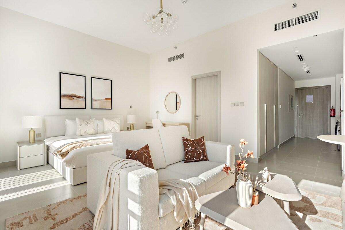 Una sala moderna con sofá, una mesita de centro y una cama con cabecera. Hay dos cuadros enmarcados en la pared, un espejo redondo y una puerta que da a otra habitación.
