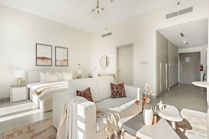 A modern living room with a sofa, a coffee table, and a bed with a headboard. There are two framed pictures on the wall, a round mirror, and a door leading to another room. - Silkhaus Al Battros (Abu Dhabi)