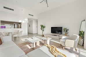A modern living room with a flat-screen TV, a dining area with a kitchen in the background, and a large window allowing natural light. - Silkhaus Al Battros (Abu Dhabi)