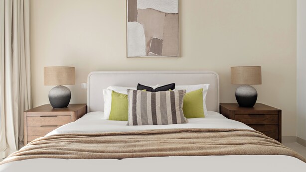 A neatly made bed with a white headboard, two bedside tables with lamps, and a large abstract painting on the wall. - Silkhaus Myrtle 1 (Dubai)
