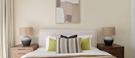 Una cama bien hecha con cabecera blanca, dos mesitas de noche con lámparas y un cuadro abstracto grande en la pared.