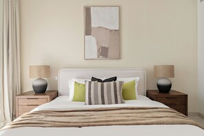 A neatly made bed with a white headboard, two bedside tables with lamps, and a large abstract painting on the wall. - Silkhaus Myrtle 1 (Dubai)