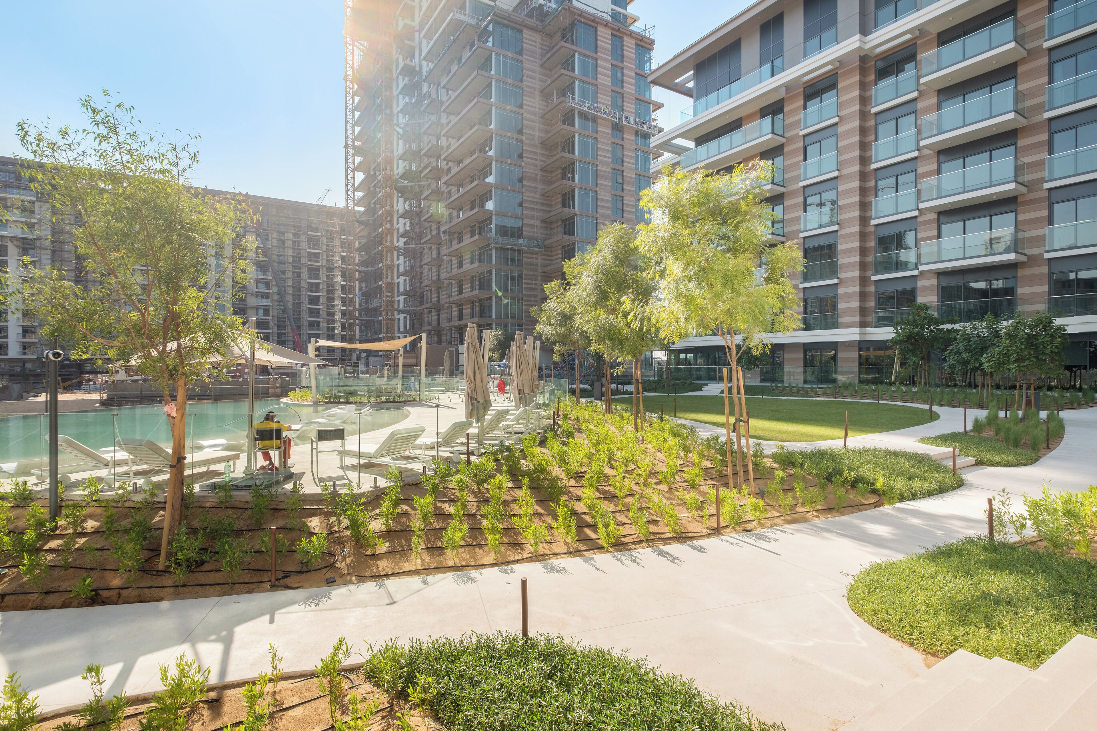 A modern outdoor pool area with lounge chairs, surrounded by greenery and adjacent to high-rise buildings.