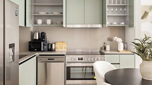 A modern kitchen with stainless steel appliances, including a refrigerator, microwave, and oven. There are open cabinets with dishes and cups, and a dining area with a table and chairs. - Silkhaus Myrtle 1 (Dubai)