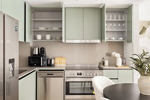 A modern kitchen with stainless steel appliances, including a refrigerator, microwave, and oven. There are open cabinets with dishes and cups, and a dining area with a table and chairs. - Silkhaus Myrtle 1 (Dubai)