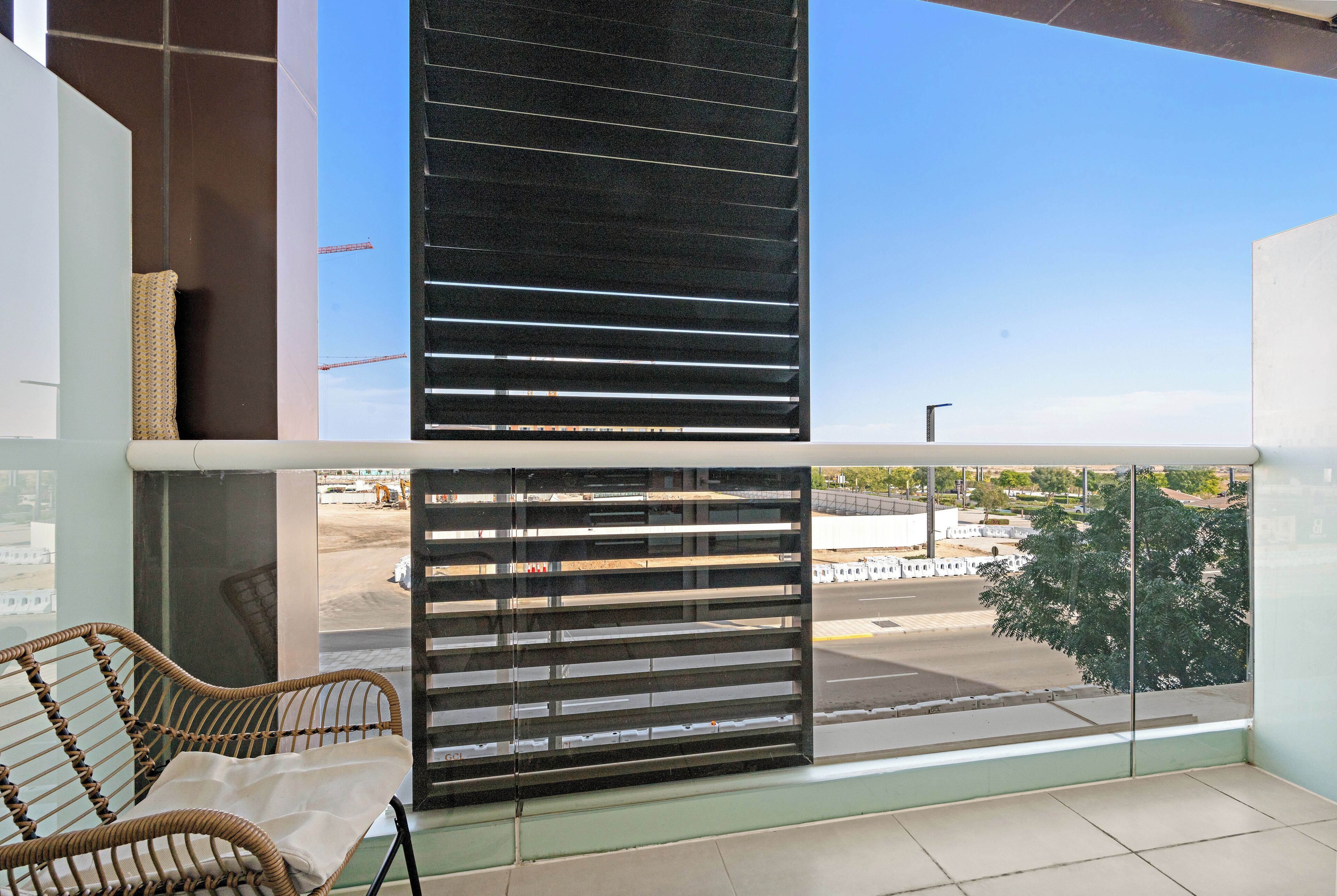 Un balcón con vista a una obra en construcción, una silla de mimbre y un panel de vidrio.
